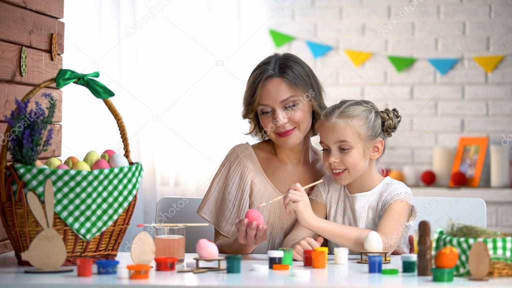 Little girl trying to paint Easter egg, loving mother helping and supporting kid