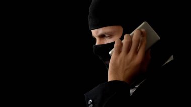 Killer in balaclava talking on phone, planning crime with partners, corruption clipart