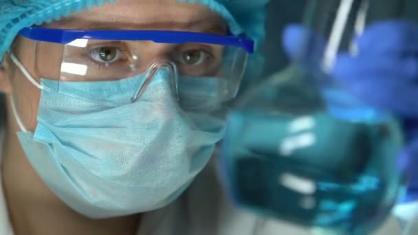 青い透明な液体が付いているフラスコを握る実験室の専門家、質を点検する — ストック動画