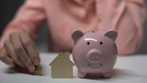Vrouwelijke hand munt aanbrengend spaarpot, houten speelgoed huis staand in de buurt van, hypotheek — Stockvideo