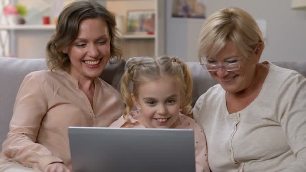 Маленькая девочка показывает смешное видео на ноутбуке маме и бабушке, юмористическое шоу — стоковое видео