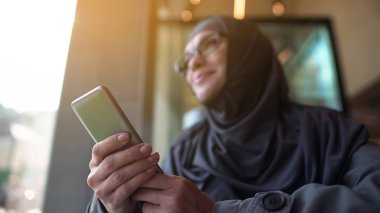 Telefon elinde tutarak, dışarıda kafe pencereden bakarak neşeli Müslüman hanım