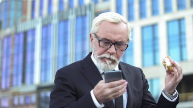 Kontrol e-posta, stresli iş, zaman eksikliği burger yemek takım elbiseli yaşlı erkek