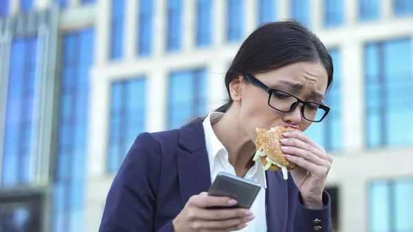 年轻的女经理吃不健康的汉堡在匆忙中 在电话里查看新闻 — 图库照片
