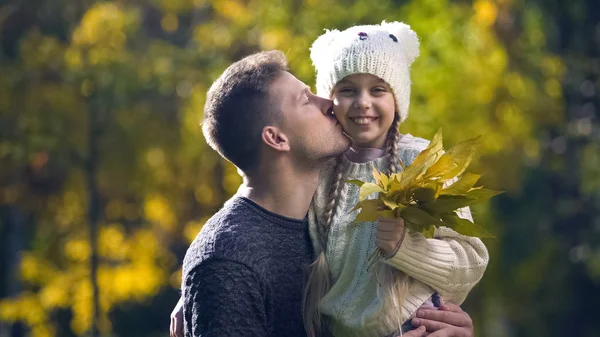 Dad holding daughter in funny hat and kissing, leisure together, autumn stroll