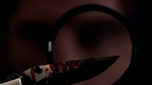Детектив смотрит на нож с кровью через увеличительное стекло, улики преступления
