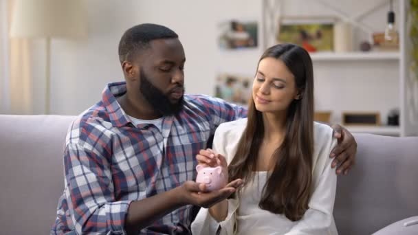 Pasangan ras campuran menempatkan koin di celengan, program sosial bagi keluarga muda — Stok Video