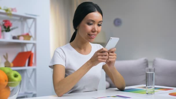 Attraente femminile sorridente che chatta con l'applicazione smartphone, seduto a casa tavolo — Video Stock