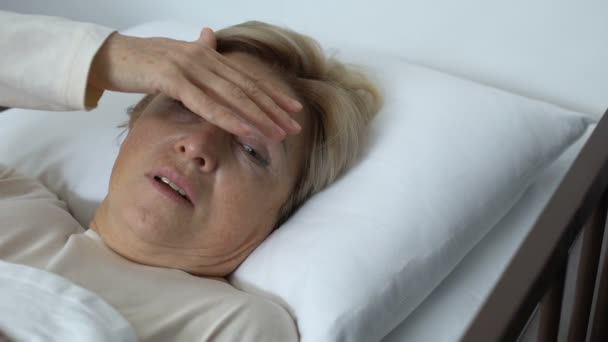 Болезненная пожилая женщина касается лба дрожащей рукой, лихорадка во время гриппа, вирус — стоковое видео