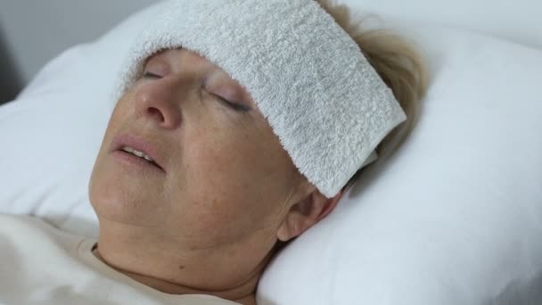 Sjuk gammal kvinna med handduk på pannan ligger i sängen, lider av kyla eller influensa — Stockvideo