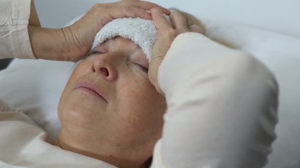Больная пожилая женщина, держащая голову с мокрым полотенцем на лбу, страдает от лихорадки — стоковое видео