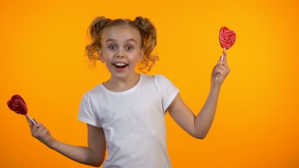 Adorable chica cerrando los ojos con piruletas en forma de corazón, sonriendo y divirtiéndose — Vídeos de Stock