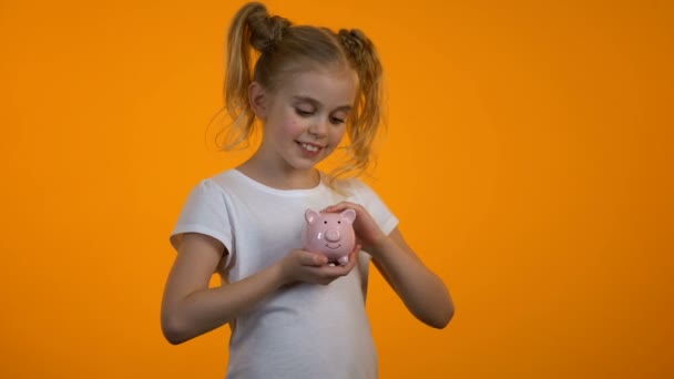 Sonriente chica poniendo moneda en la hucha y mirando a la cámara, ahorros para el futuro — Vídeos de Stock