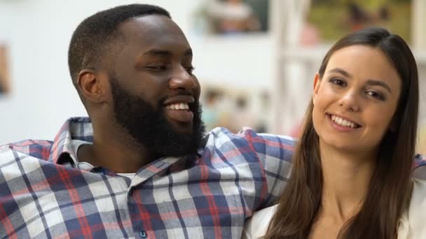 Hombre negro mirando a su bonita novia, pareja sentada en el sofá y abrazándose — Vídeos de Stock