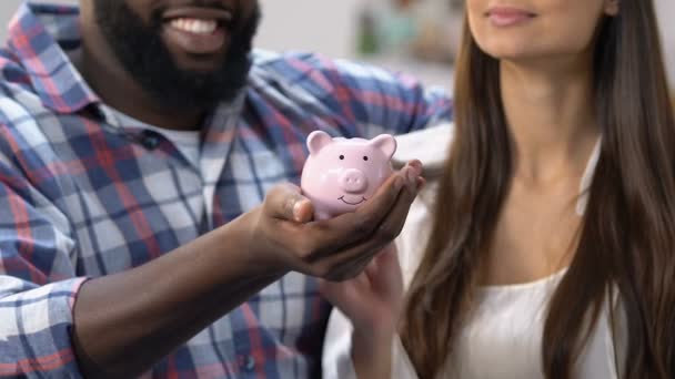Африканский американец держит копилку, женщина кладет монету внутрь, бюджет — стоковое видео