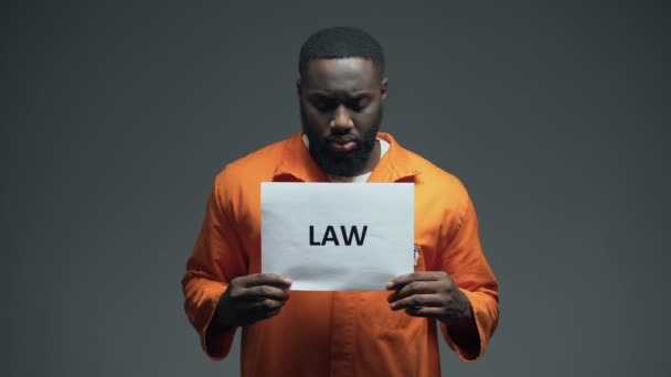 Homem negro preso segurando sinal de lei na cela, olhando para câmera, direitos humanos — Vídeo de Stock