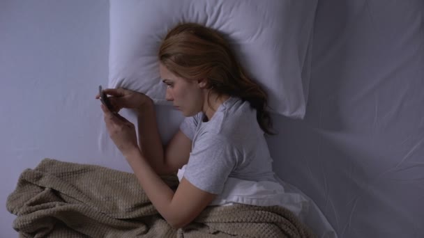 ベッドに横たわるスマートフォンで古いメッセージを読んで泣く悲しい女性、思い出 — ストック動画