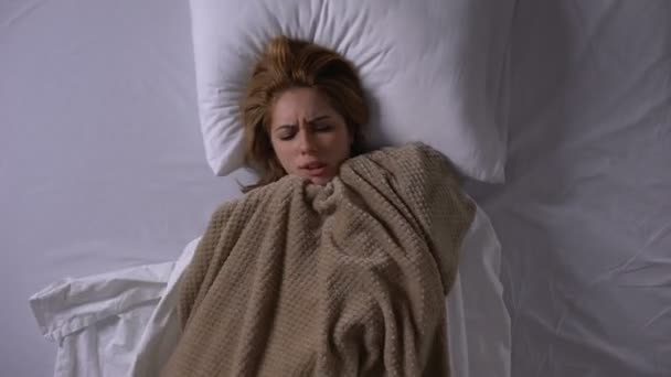 Женщина, покрытая одеялом, лежит в постели, чувствует лихорадку, симптомы простуды — стоковое видео