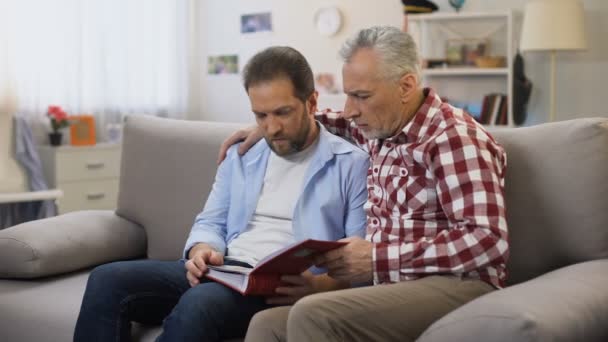 Hombres adultos pensativos sosteniendo álbum y viendo fotos, recordando a sus parientes — Vídeo de stock