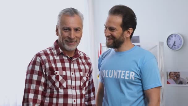 若い男性ボランティア抱擁シニア男性と両方のカメラに笑顔,慈善団体 — ストック動画