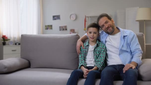 Mutlu gülümseyen baba ve okul çocuğu oğlu kanepede oturan, kamera, aile bakıyor — Stok video