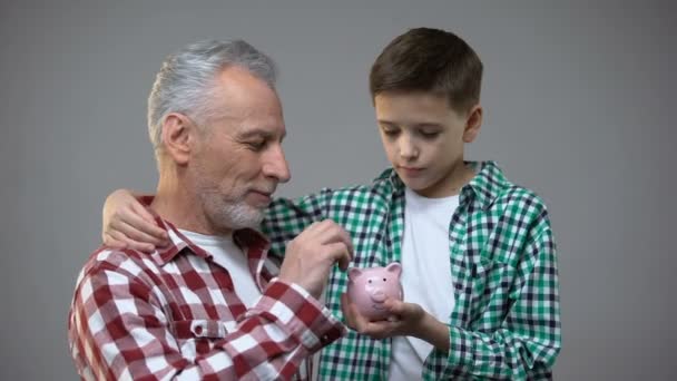 Ηλικιωμένος άνθρωπος βάζοντας κέρματα σε μικρό αγόρι κουμπαράς, εξοικονόμηση για το μέλλον, τραπεζικές — Αρχείο Βίντεο