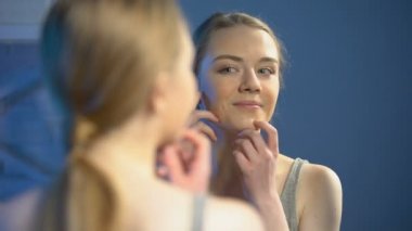 Mutlu genç kadın ayna yansıması bakarak, cilt tedavisi memnun