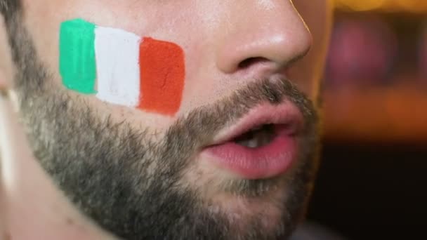 Ventilador masculino com bandeira italiana na bochecha emocionalmente torcendo para a equipe esportiva nacional — Vídeo de Stock