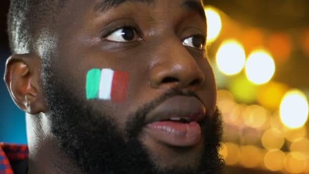 Fanático de los deportes afroamericanos regocijando victoria del equipo favorito, bandera italiana en la mejilla — Vídeos de Stock