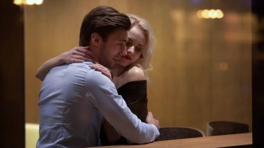 Sarılıp okşayan genç bir adam, güzel sarışını, akşam vakti dinlenme odasında.