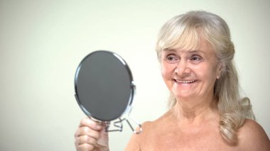 Yaşlı kadın aynaya bakıyor, kozmetik işlemlerden sonra taze cilt seviniyor