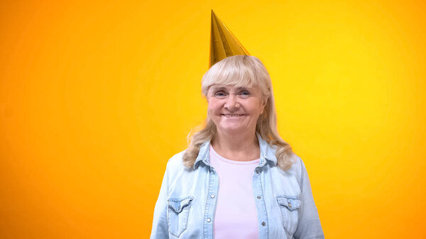 Positive elderly lady in party hat smiling to camera, birthday celebration