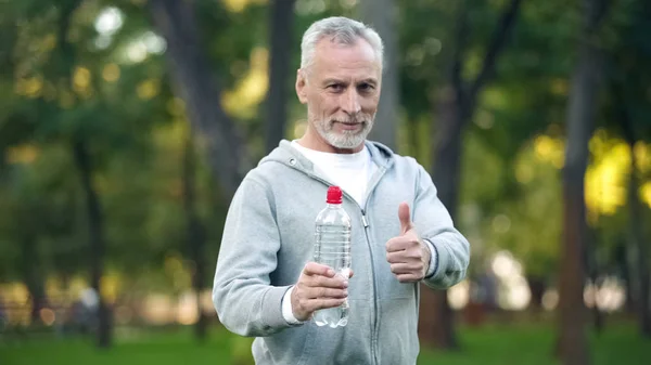 ボトル入り飲料水と親指 アクア バランスを示すスポーツ ウエアで年配の男性 — ストック写真