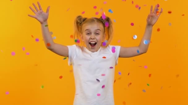 Pequeña chica emocionada sonriendo de pie bajo la lluvia de confeti sobre fondo naranja — Vídeos de Stock