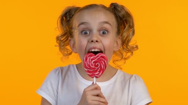 Aranyos lány Pigtailek nyalás szív alakú cukorka, elkülönített sárga háttér — Stock videók