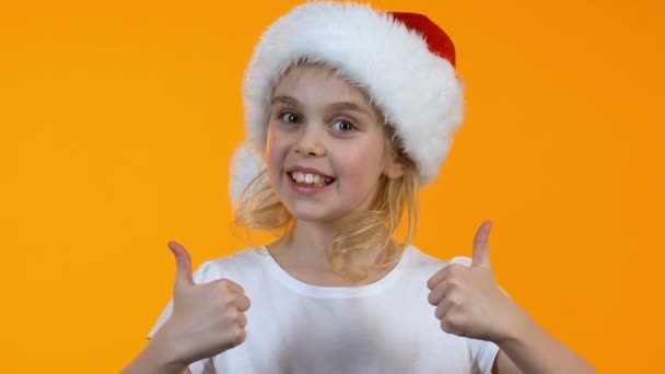 Linda niña en Santa Claus sombrero mostrando los pulgares hacia arriba y sonriendo en la cámara — Vídeo de stock