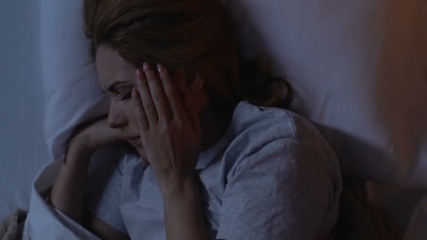 Плаче жінка лежить в ліжку вночі, тримає голову, страждає від мігрені — стокове відео