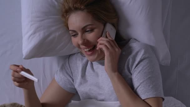 Улыбающаяся женщина в постели проводит тест на беременность и говорит по телефону, положительный результат — стоковое видео