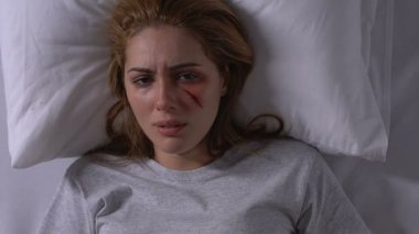 Yaralı yüzüne dokunan ve yatakta ağlayan kadın, aile içi şiddet kurbanı, umutsuzluk