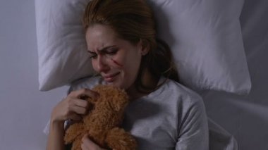 Yaralı yüzü oyuncak ayıya sarılan, yatakta ağlayan, kurbanın desteğe ihtiyacı olan bir bayan