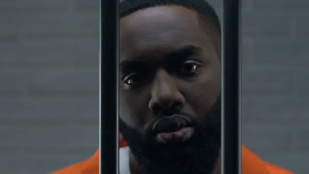 Delincuente afroamericano serio mirando a la cámara a través de los bares de la prisión, desesperación — Vídeo de stock