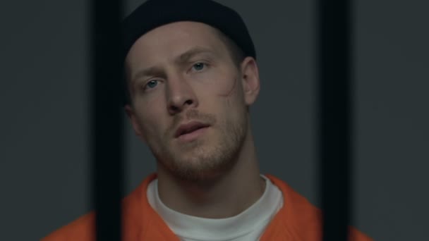 Zelfverzekerd crimineel met littekens op het gezicht kijken naar de camera achter de gevangenis bars — Stockvideo