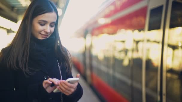 Jonge dame typen op de smartphone op het platform in de buurt van de trein en glimlachend naar camera — Stockvideo