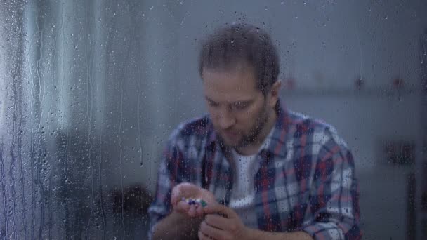 絶望的な男性は雨の窓の後ろに手のひらに錠剤を保持し、自殺未遂 — ストック動画