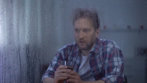 Депресивний чоловік п'є віскі один на дощовий день, думаючи про життєві проблеми — стокове відео