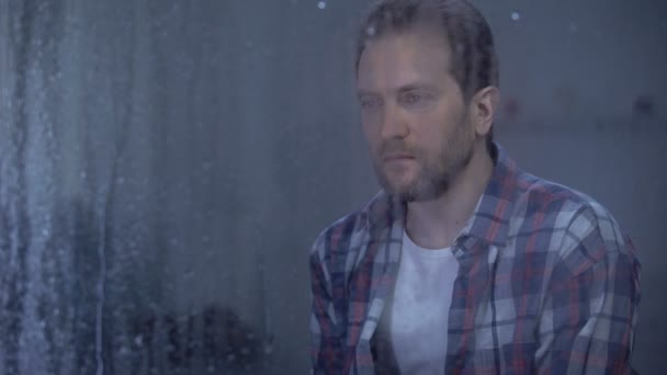 Sad man looking through window during rainy weather, thinking about life problem — Stock Video