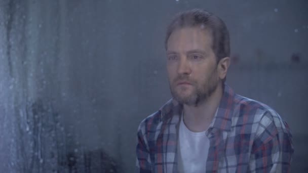 Одинокий человек смотрит в окно в дождливый день, проблемы с депрессией — стоковое видео