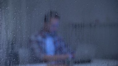 Odaklanmış orta yaşlı adam yağmurlu pencere arkasında dizüstü bilgisayar üzerinde çalışan, freelancer