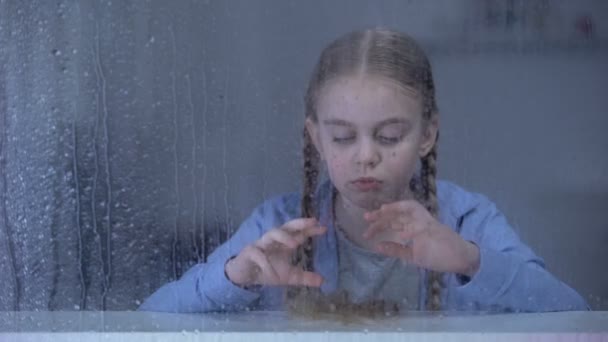 Скучная маленькая девочка смотрит в камеру через окно в дождливую погоду, одиночество — стоковое видео