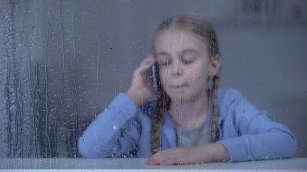 Θλιβερό μικρό κορίτσι μιλάει τηλέφωνο σε βροχερή μέρα, μένοντας στο σπίτι μόνος, αγνοούμενη μητέρα — Αρχείο Βίντεο